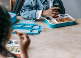 Crop faceless classmates sitting at table with lunch boxes with various food and eating during break